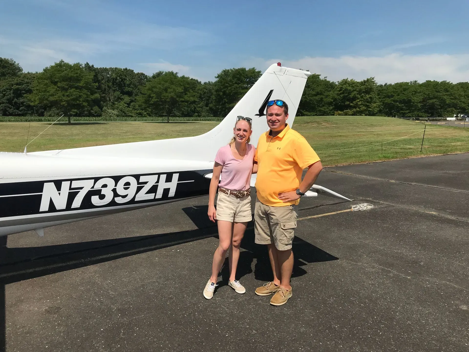 Two people standing by a small airplane, one in yellow, one in pink. Sunny day, airfield setting.