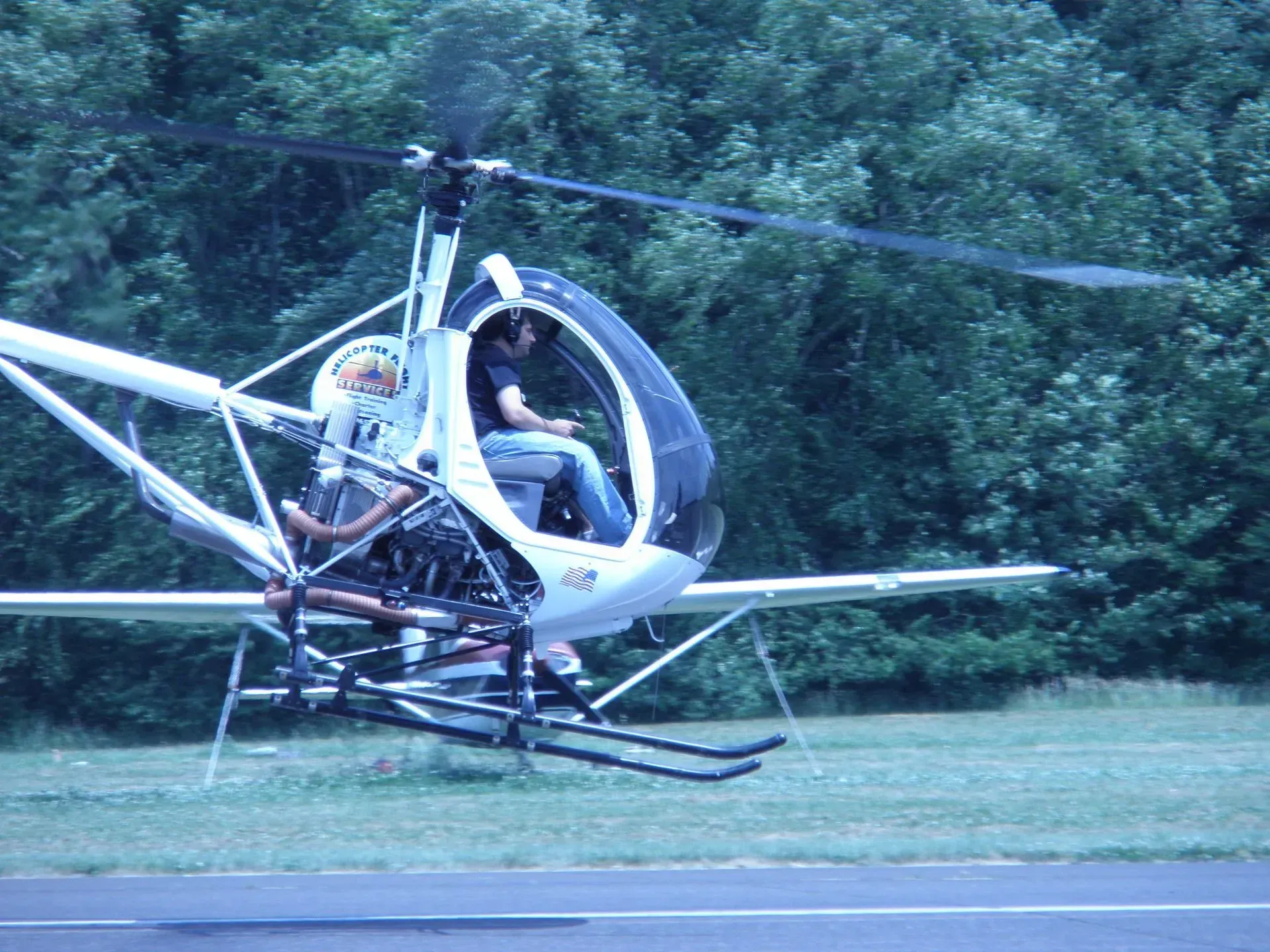 Two people in a small, open-cockpit helicopter on a grassy field, rotor blades spinning.