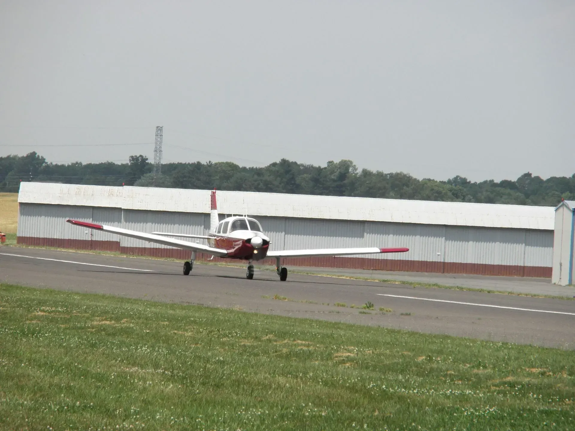 Small, red and white airplane on a runway, approaching a white hangar on a sunny day.