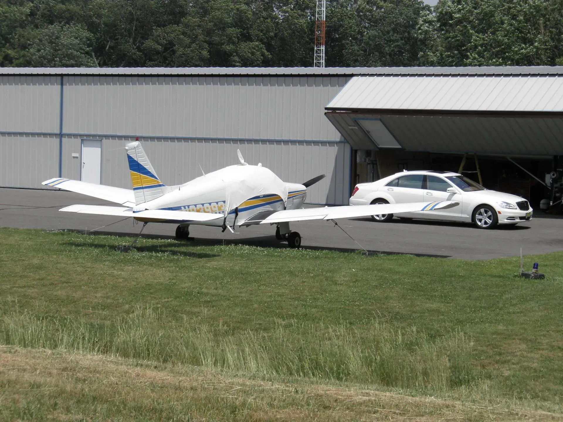 Small airplane parked on grass near a white car and a hangar.