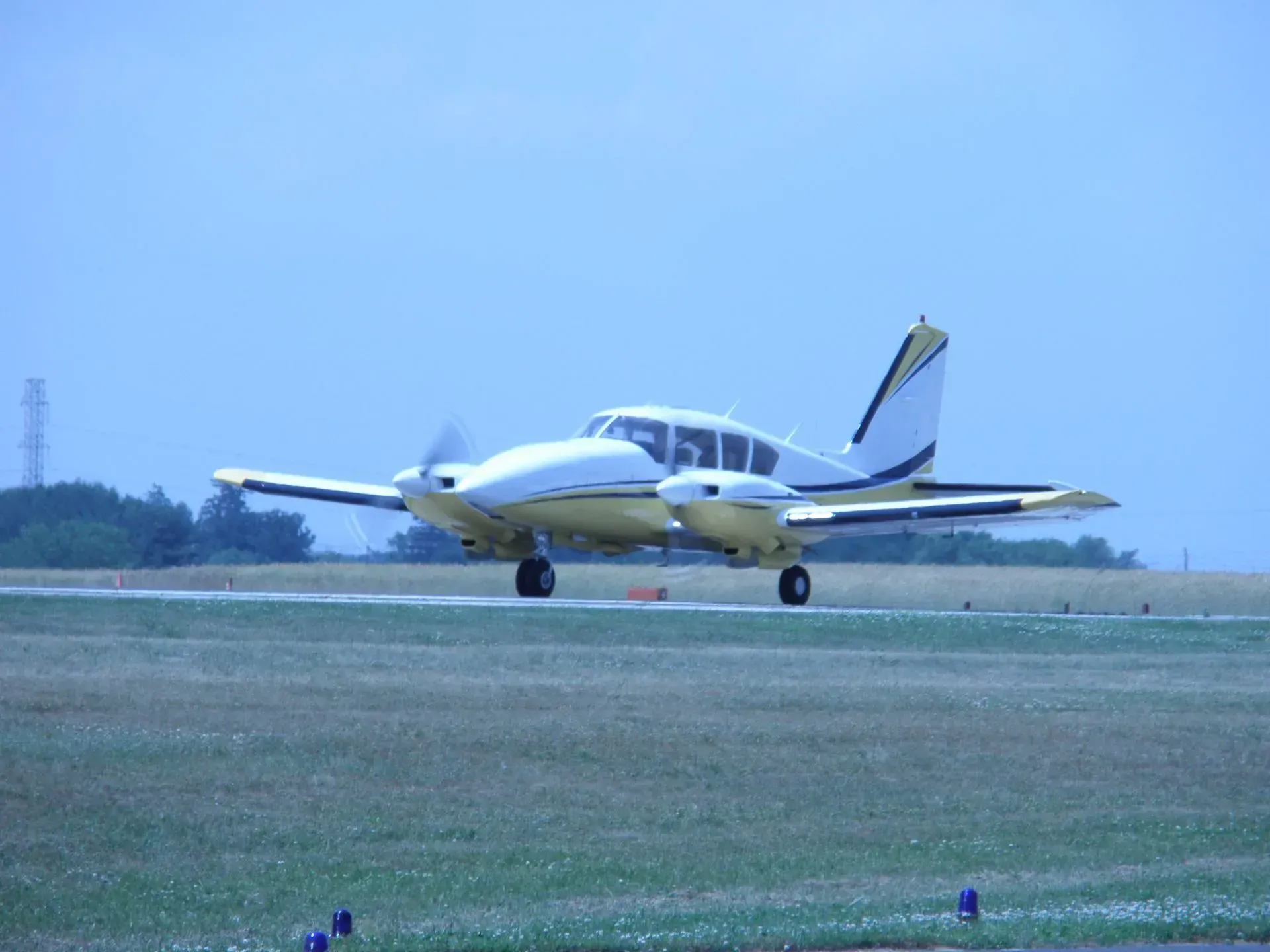 Twin-engine airplane on a runway, white and yellow paint. Propeller spinning, blue sky background.