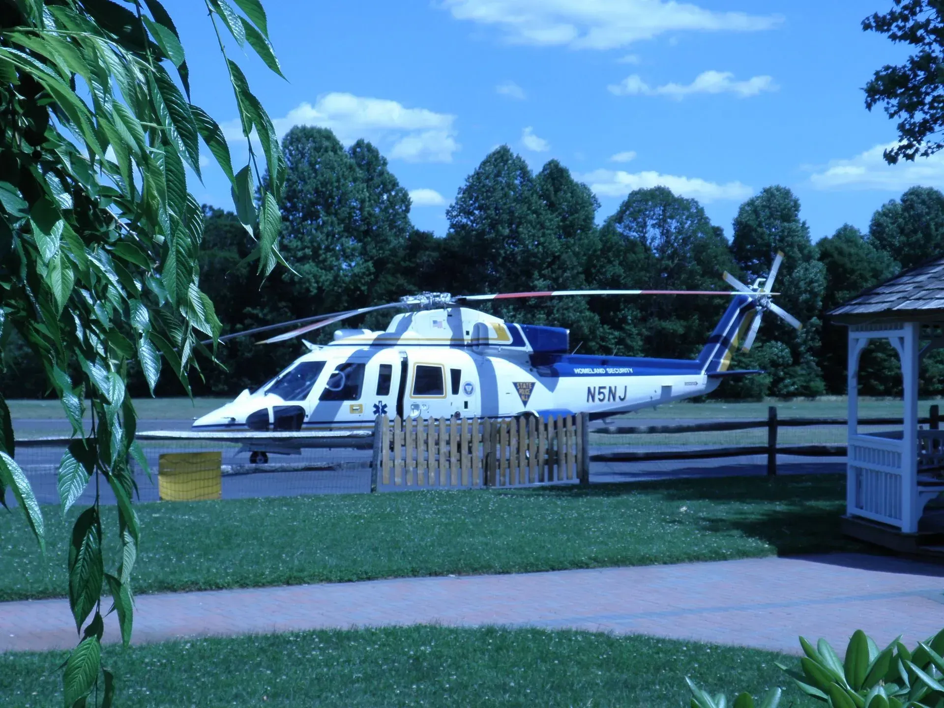 Helicopter on the ground, painted white and blue, near a wooden fence and gazebo on a sunny day.