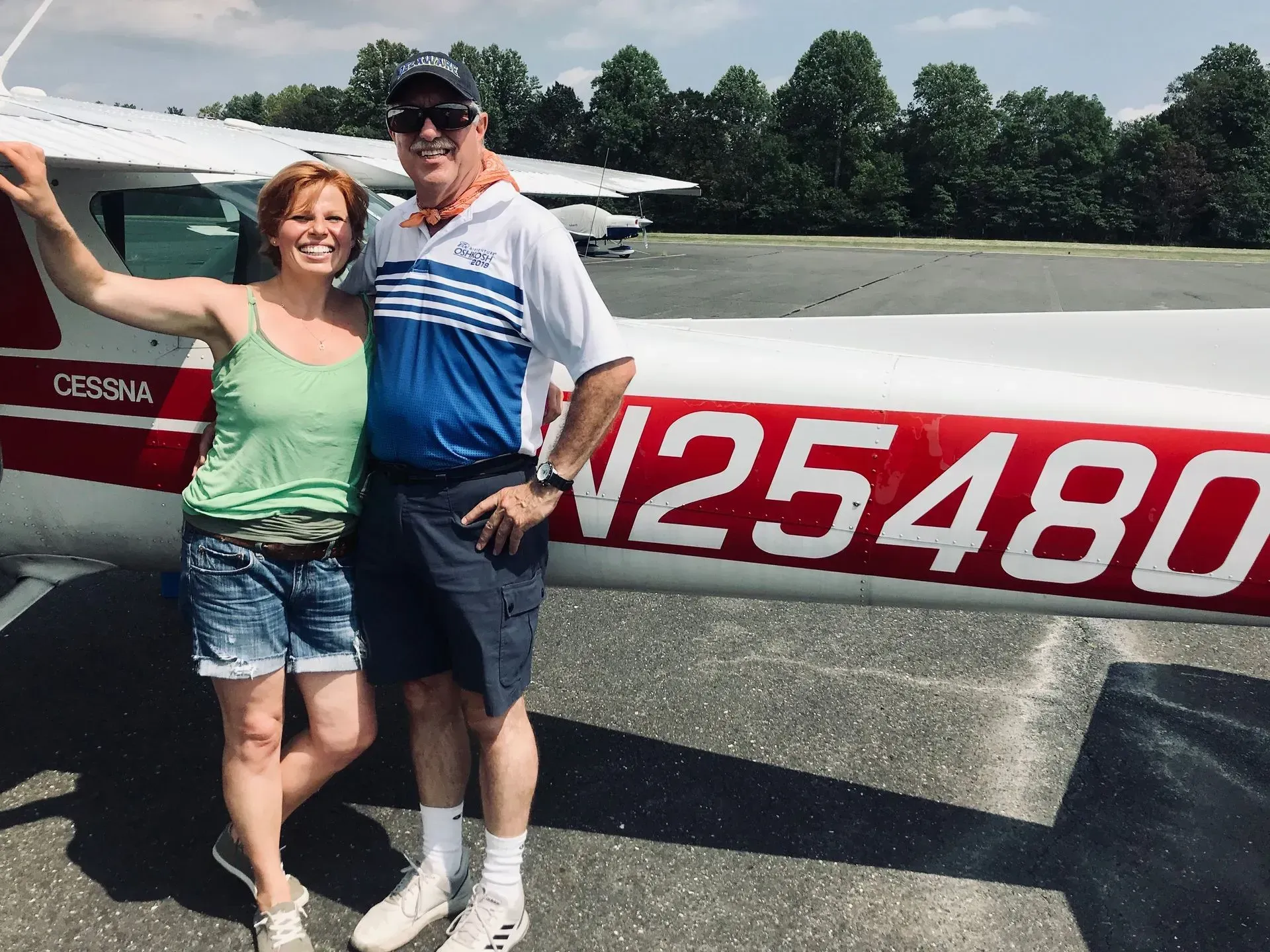 Two people pose by a Cessna airplane; woman in green top, man in blue shirt. Sunny day.