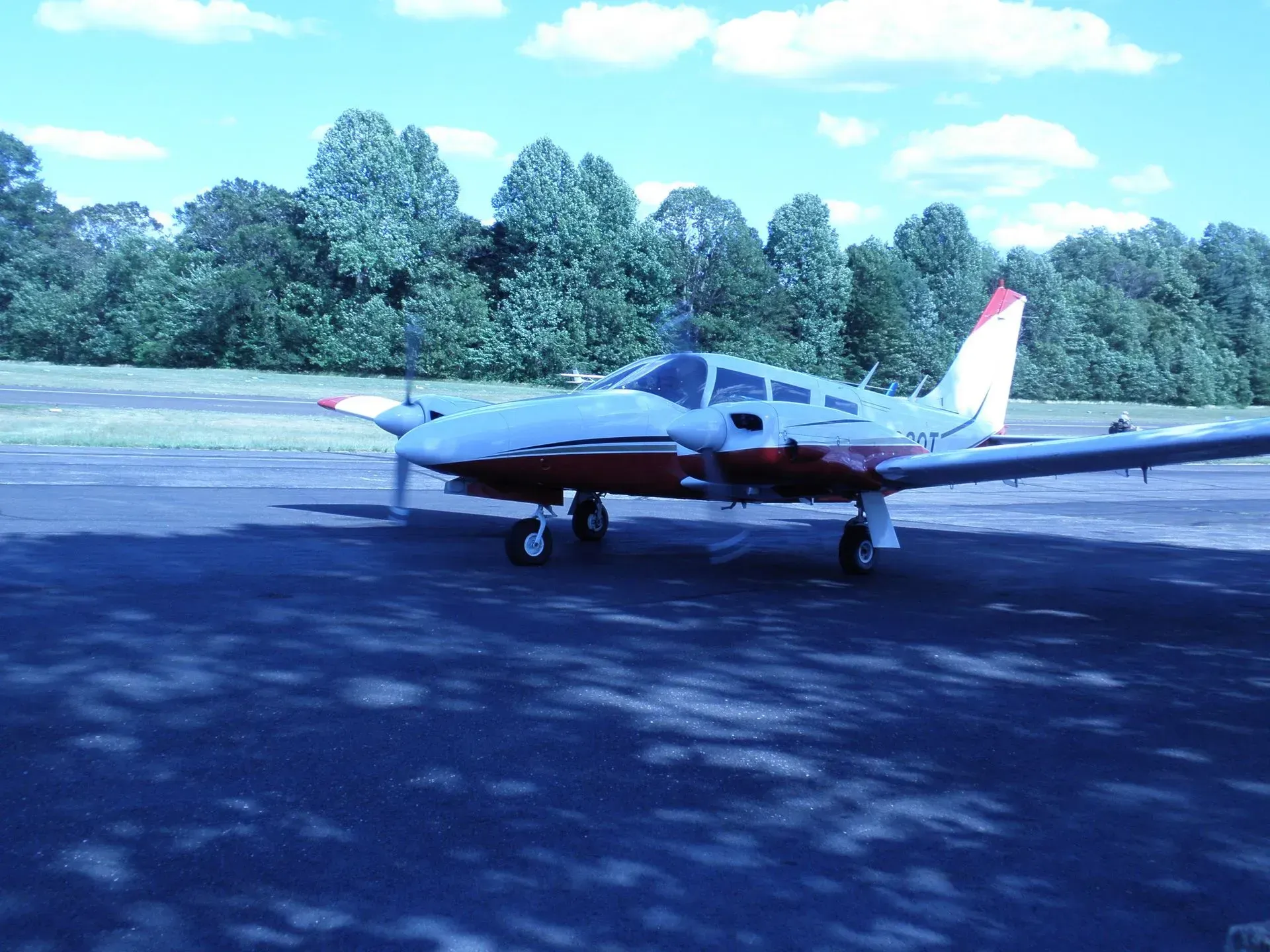 Small red and white airplane on a tarmac, trees in the background under a blue sky.