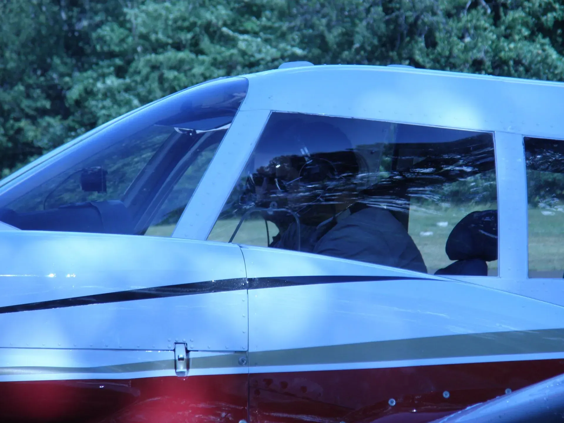 Close-up of airplane cockpit. Pilot visible inside, reflected in the window. White plane with red stripe, set against green trees.
