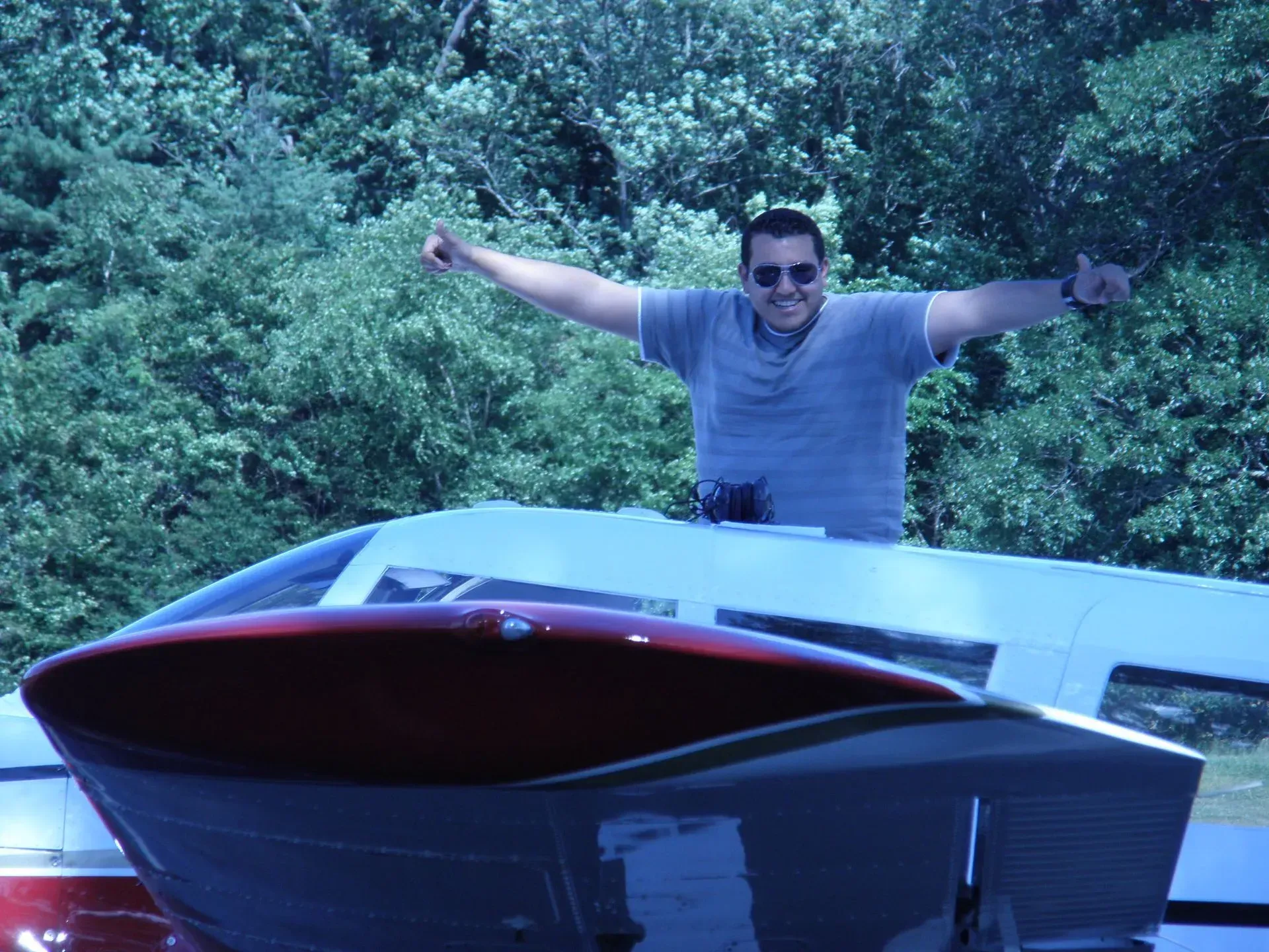 Man standing on aircraft, arms raised in thumbs up gesture; white and red plane in front of trees.