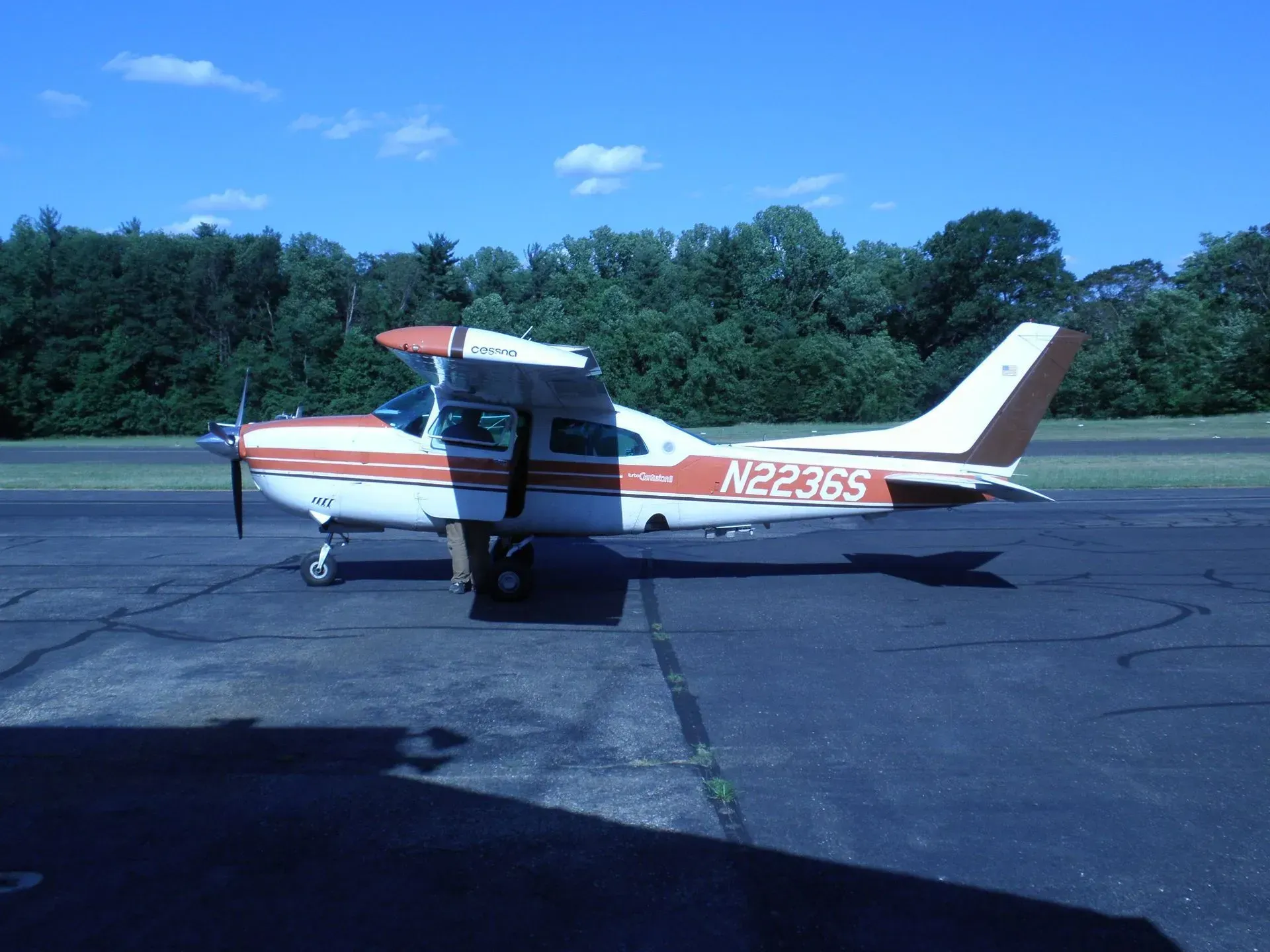 Cessna 182 airplane, white and orange, parked on a tarmac with trees in the background, sunny day.
