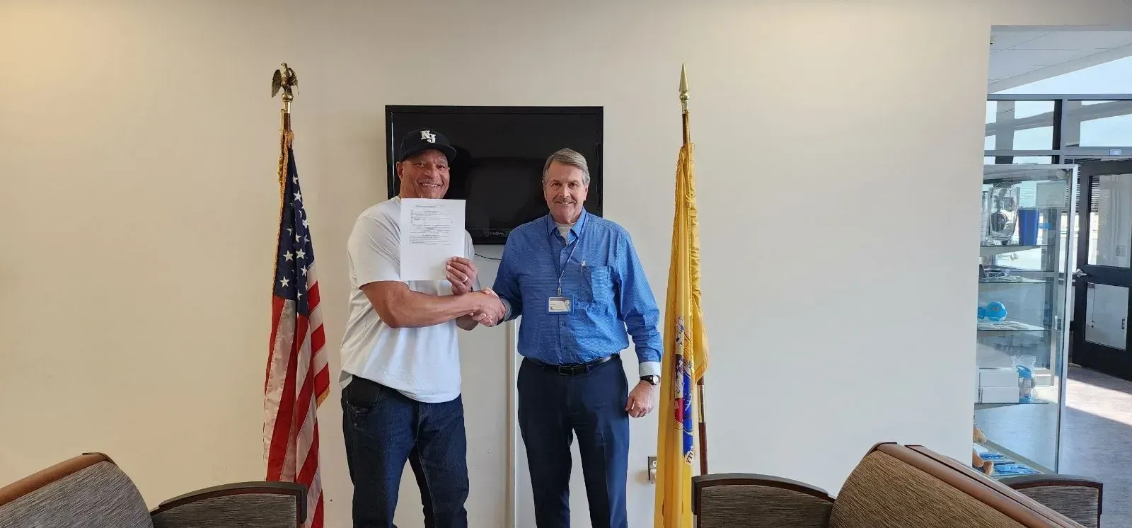 Two men shaking hands in an office, one holds a paper. US and state flags are in background.