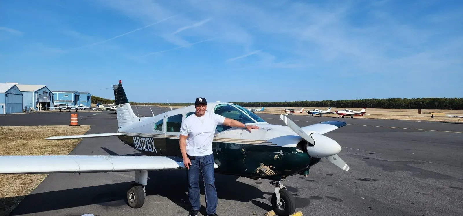 Man standing next to a small airplane on a tarmac, pointing toward the aircraft, blue sky.