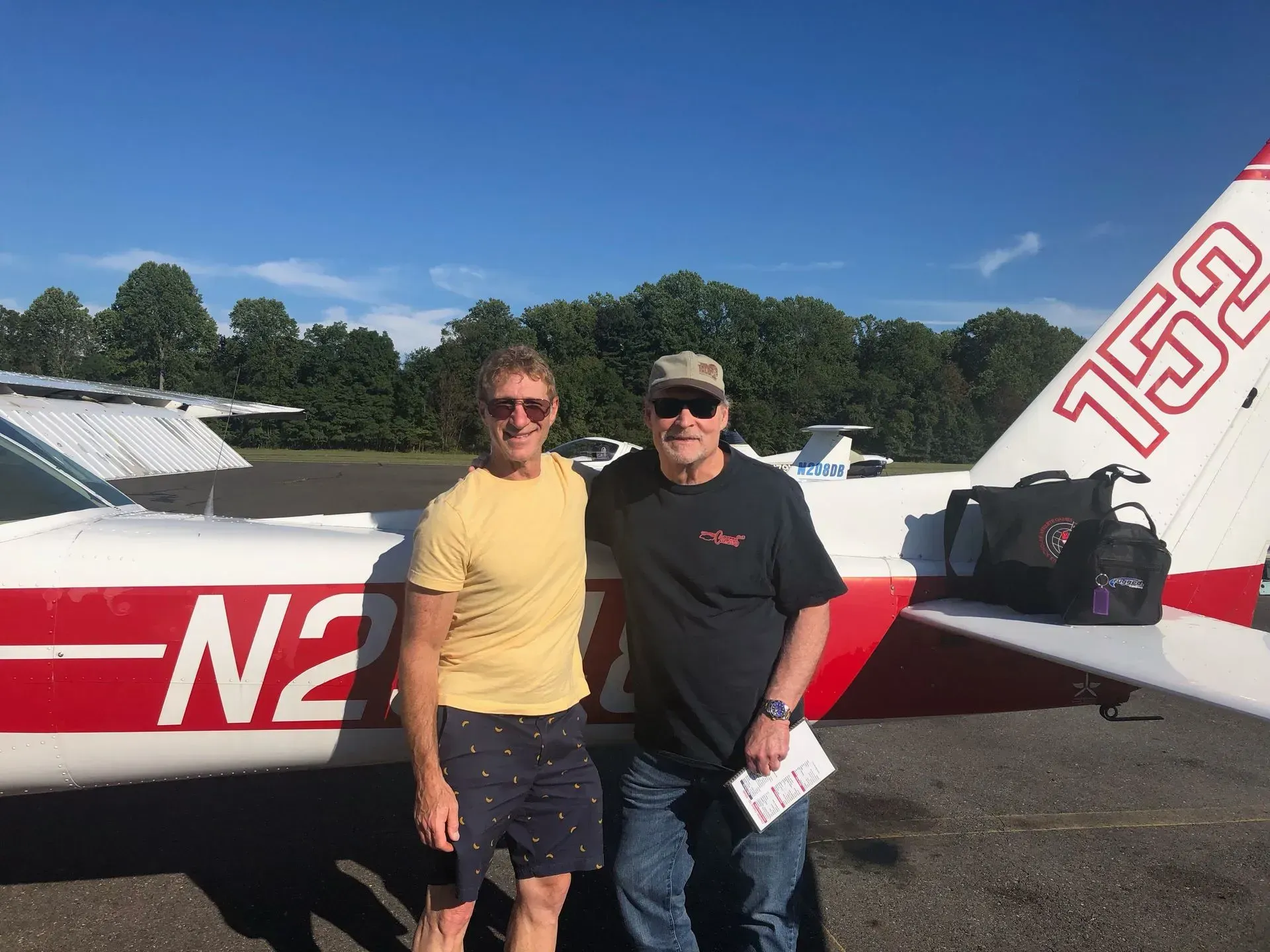 Two men stand by a small plane with red and white detailing on a sunny day. One man has his arm around the other.