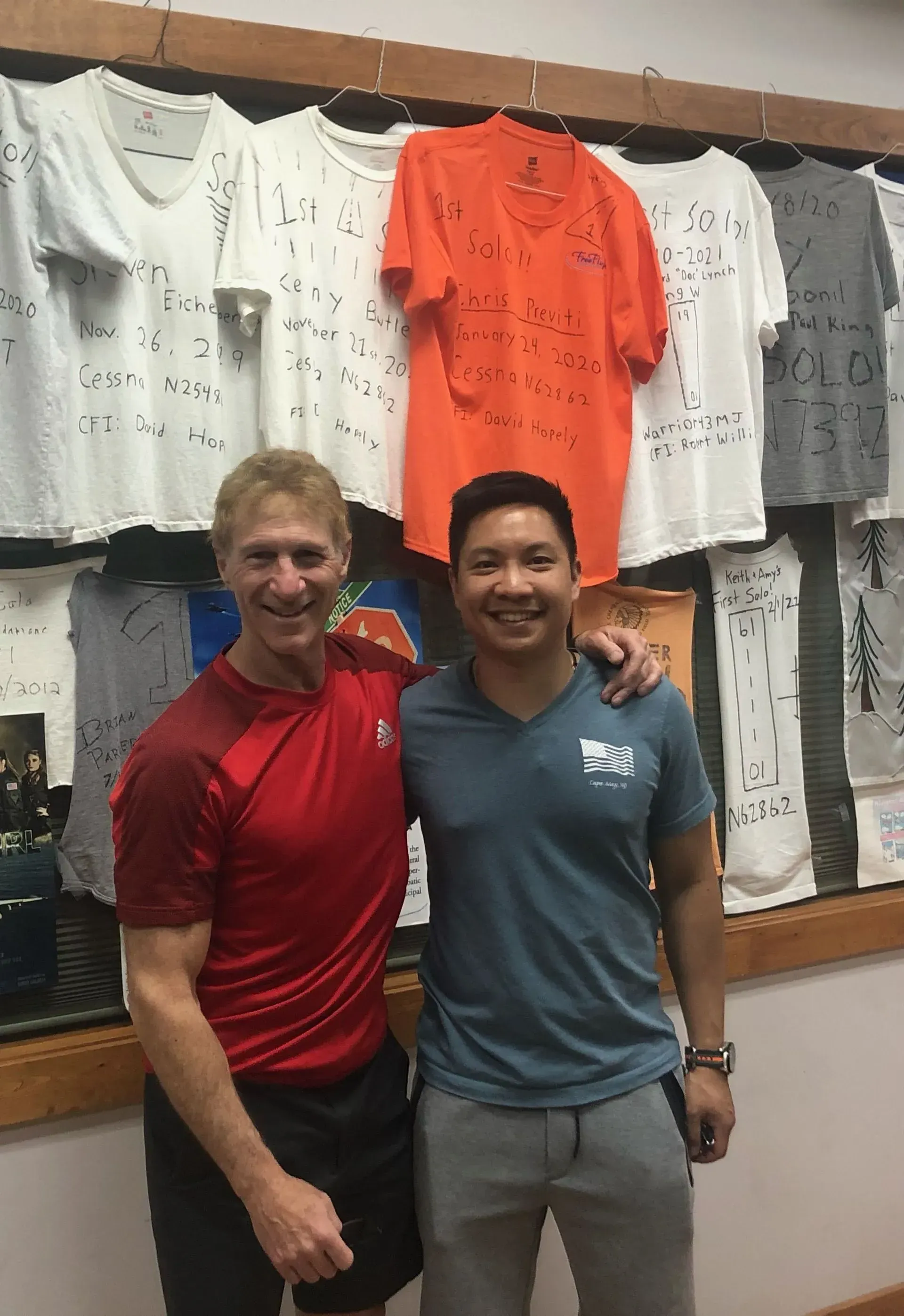 Two men pose together indoors; one in red, one in blue, with signed shirts in the background.