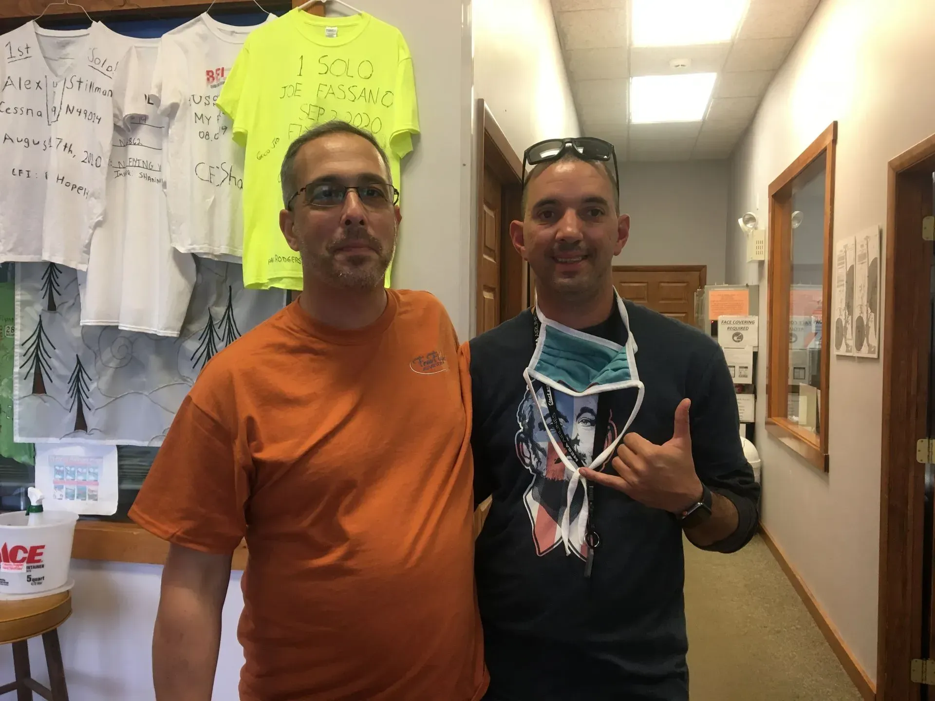 Two men smiling indoors, one wearing orange, the other with a mask around his neck, giving a shaka sign.