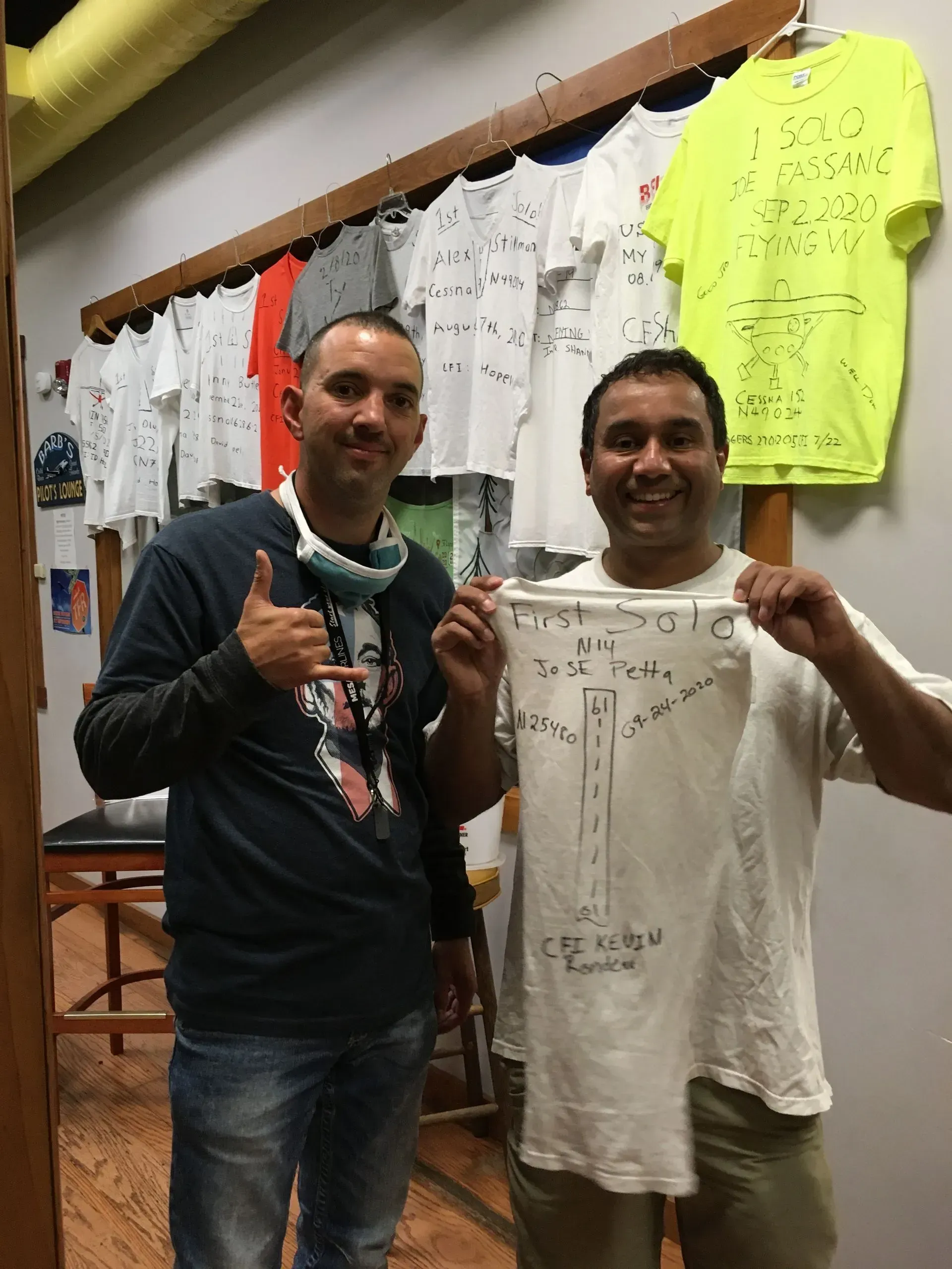 Two men holding a shirt with a drawing of a street intersection. They are indoors with white shirts hanging on a rack.