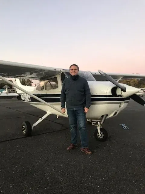Man standing by a small airplane on a tarmac. He’s smiling, wearing a blue sweater and jeans. Dusk setting.