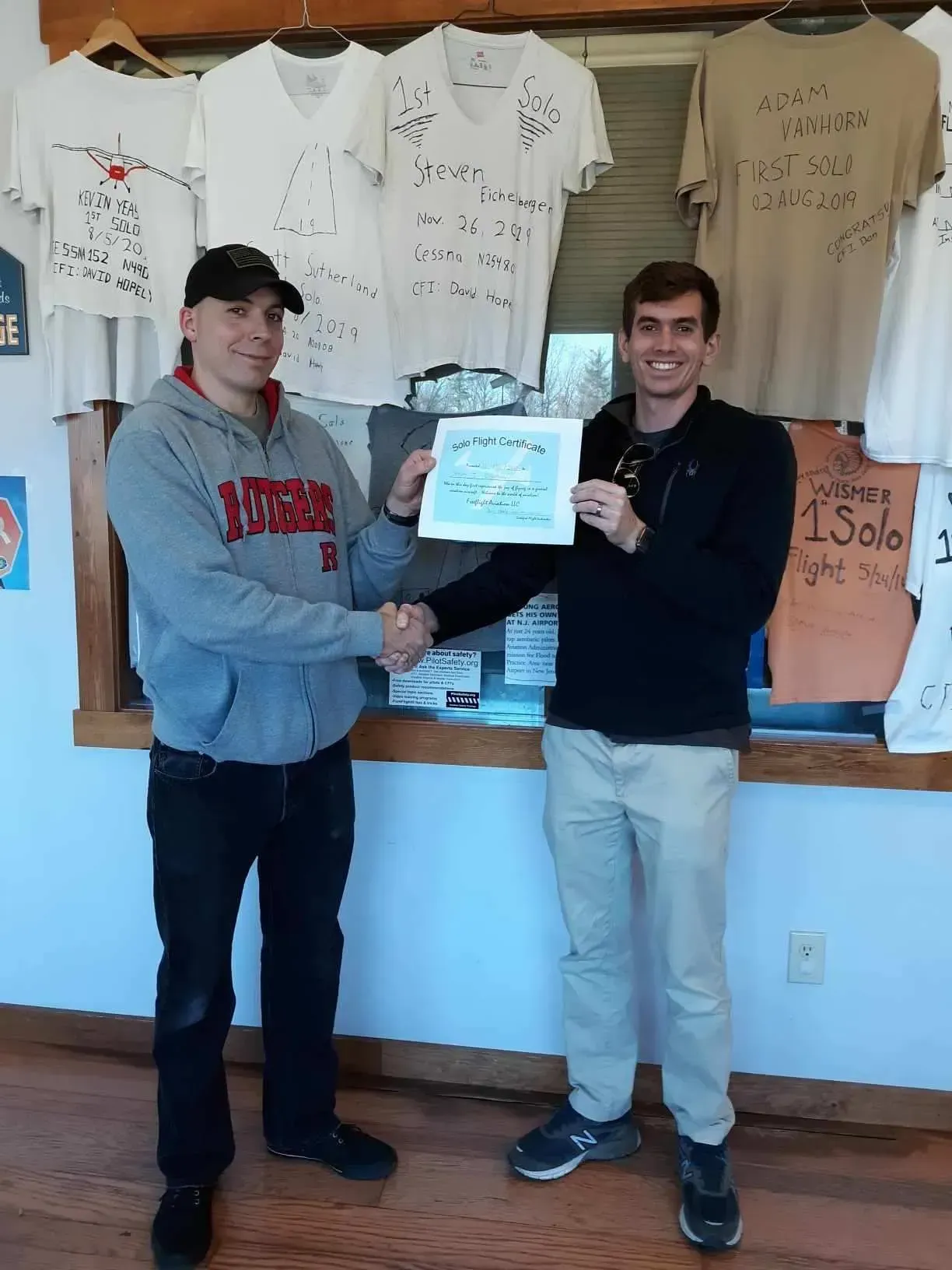 Two men shake hands, one holding a framed certificate. T-shirts hang on the wall behind them.