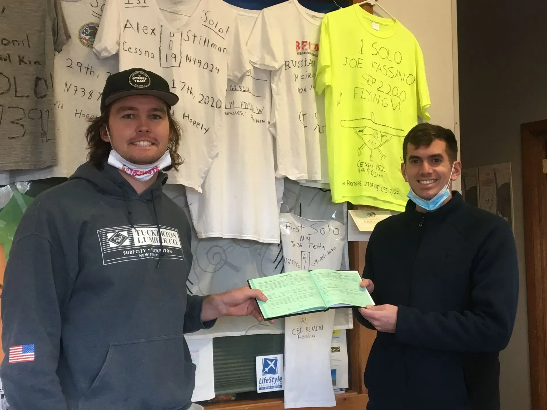Two men holding a green check, standing in front of signed t-shirts. One man is in a hoodie and baseball cap, the other in a sweater.