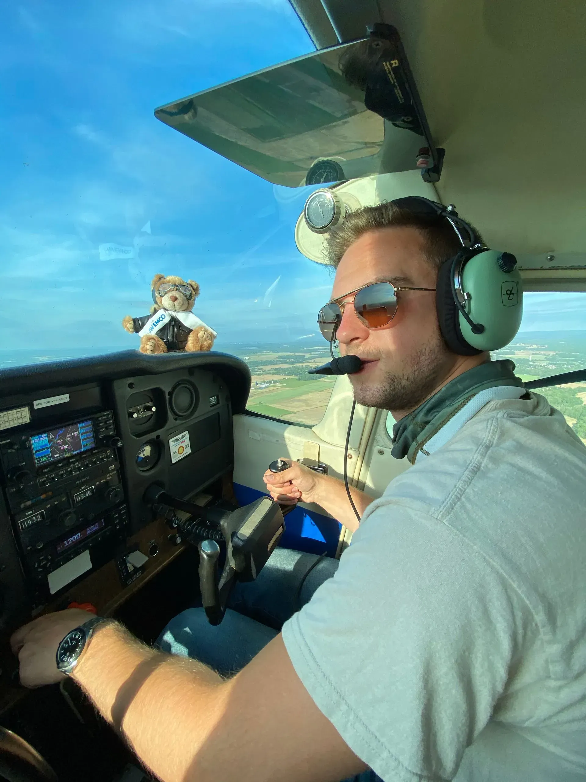 Man in airplane cockpit, wearing headset and sunglasses, looking out. A teddy bear sits on the dashboard.