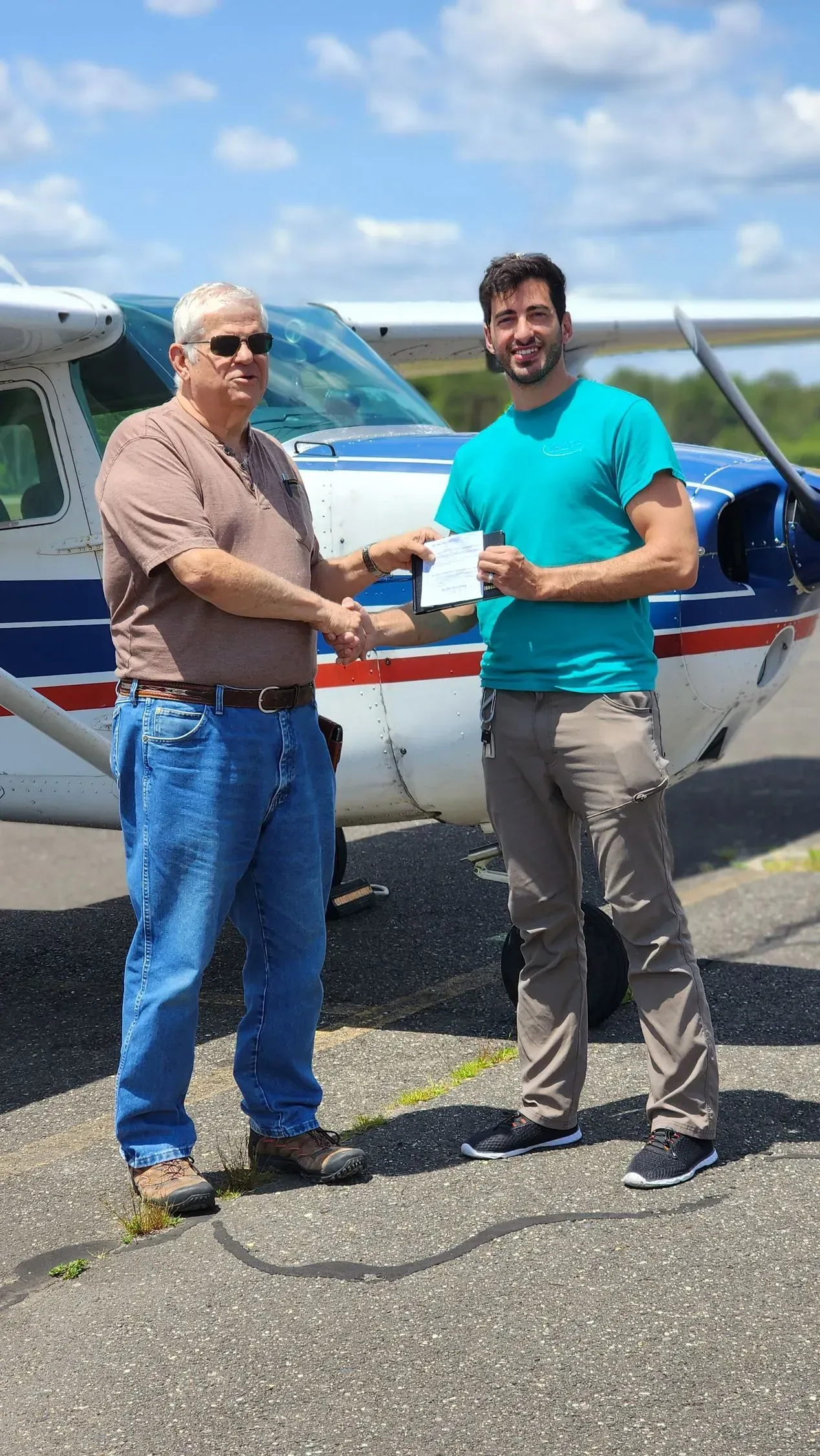Man in brown shirt hands check to man in teal shirt near airplane, outdoors.