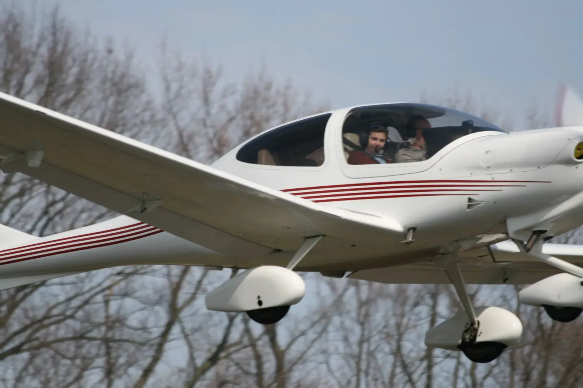 White airplane with red stripes in flight, two people in the cockpit, trees in the background.