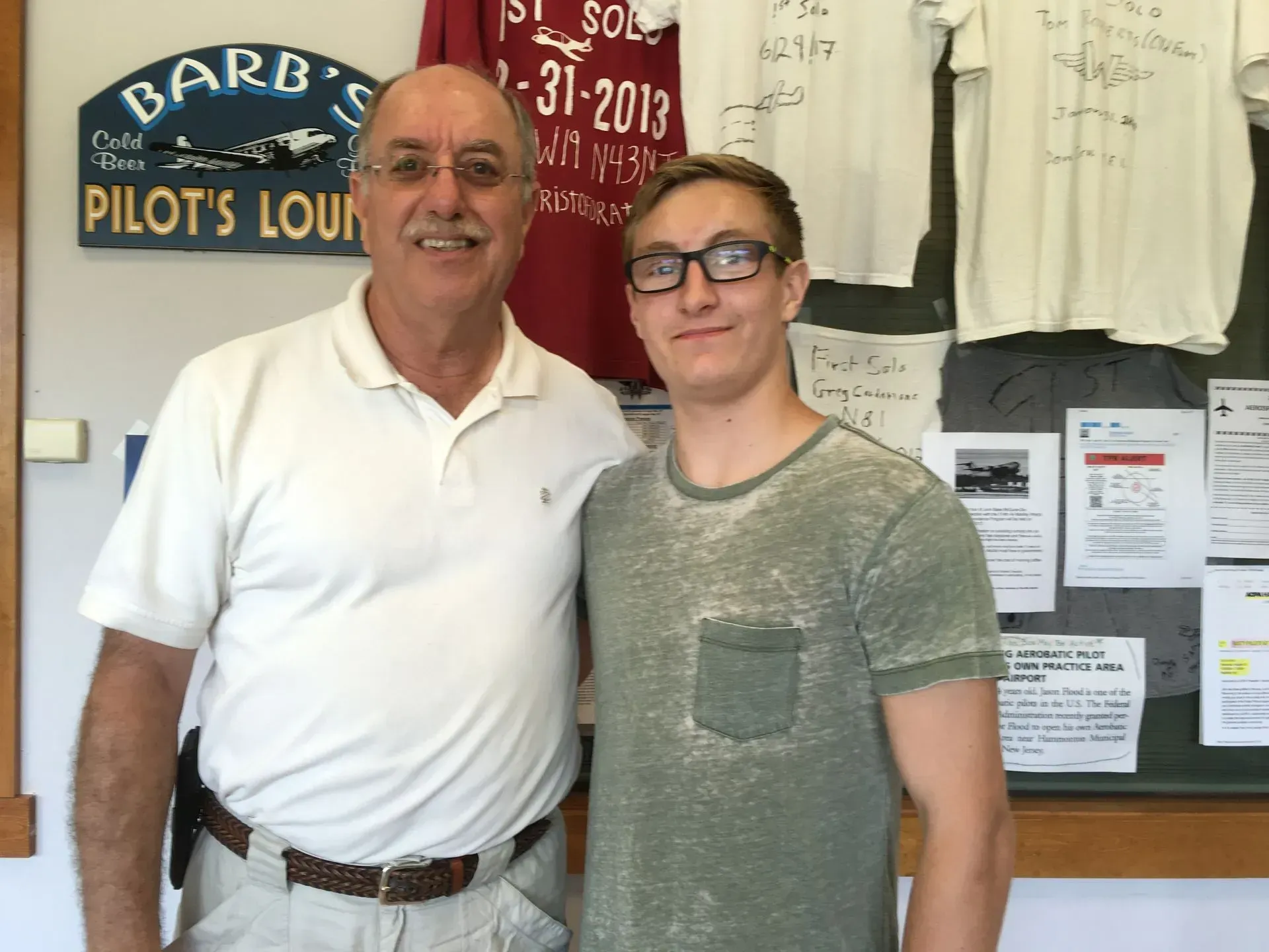 Two men posing for a photo inside a pilot's lounge. One man in a white shirt and belt, the other in a green t-shirt and glasses.