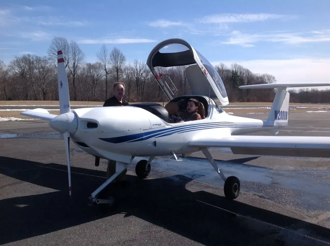 A white single-engine plane on a runway, open canopy, two people inside, one person standing beside it.
