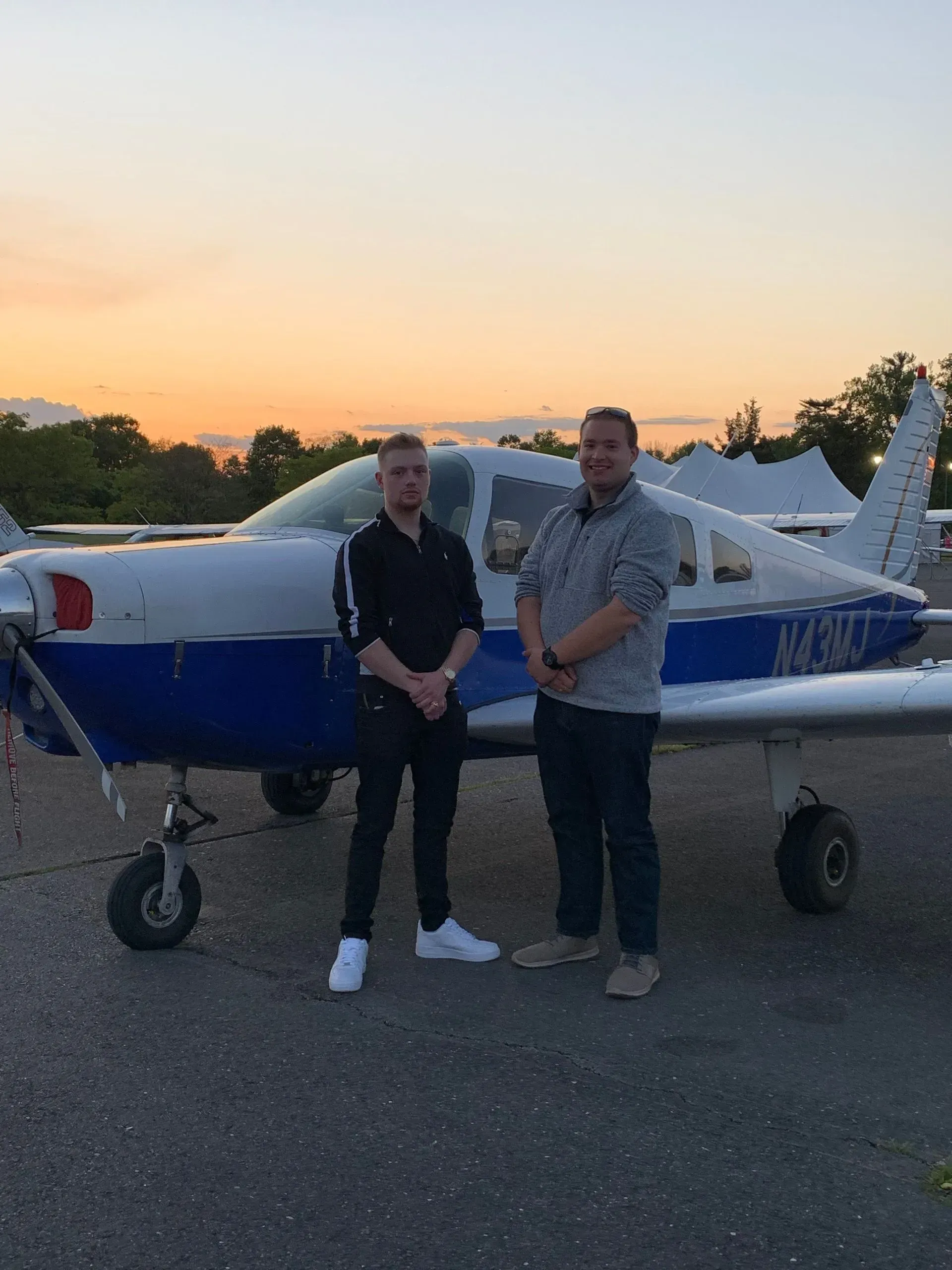 Two men stand by a small blue and white airplane at dusk.