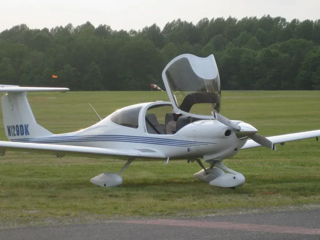 White Diamond aircraft on a grassy field with the canopy open, near trees.