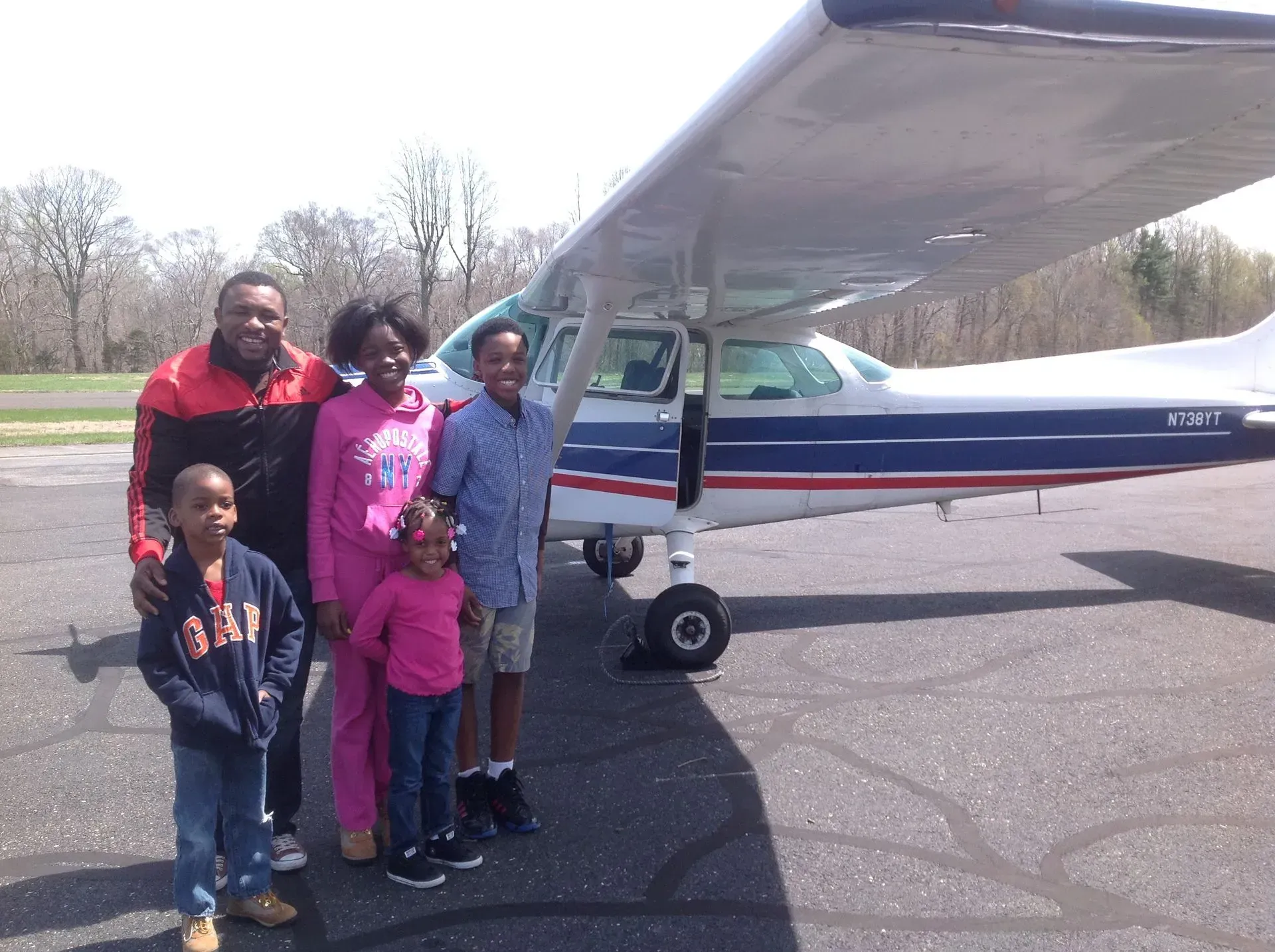 Family poses next to a small airplane on a tarmac, sunny day.