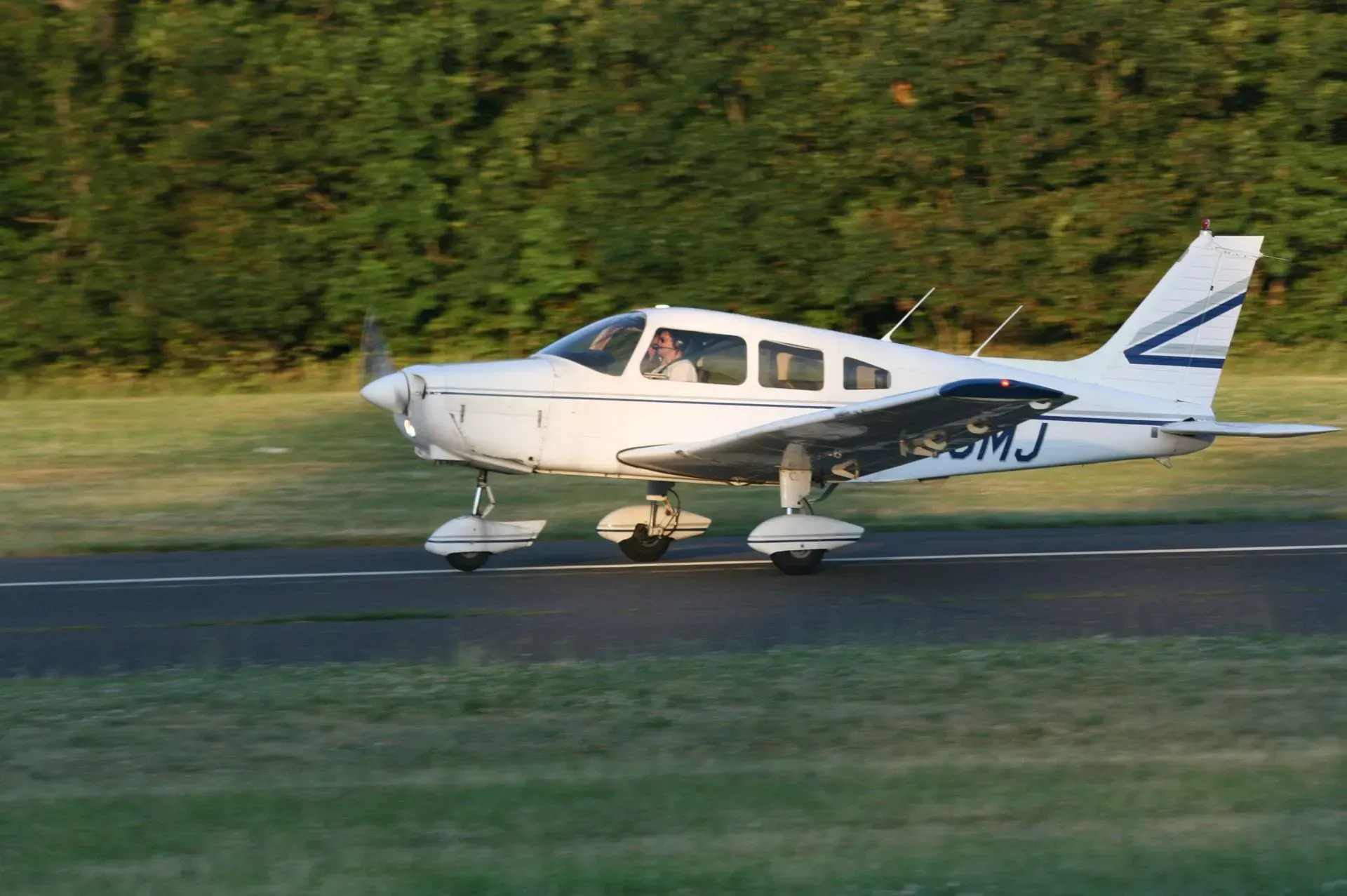 White airplane on a runway, preparing for takeoff. Pilot visible. Green trees in background.