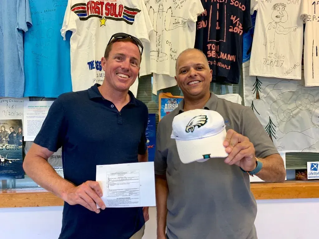 Two men smiling, one holding a paper, the other an Eagles hat, in front of shirts and a wall.