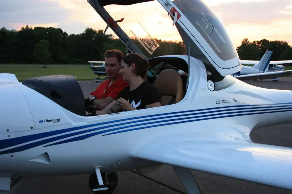 Two people in a small white airplane cockpit. One is wearing an orange shirt, and the other a black shirt. Dusk setting.