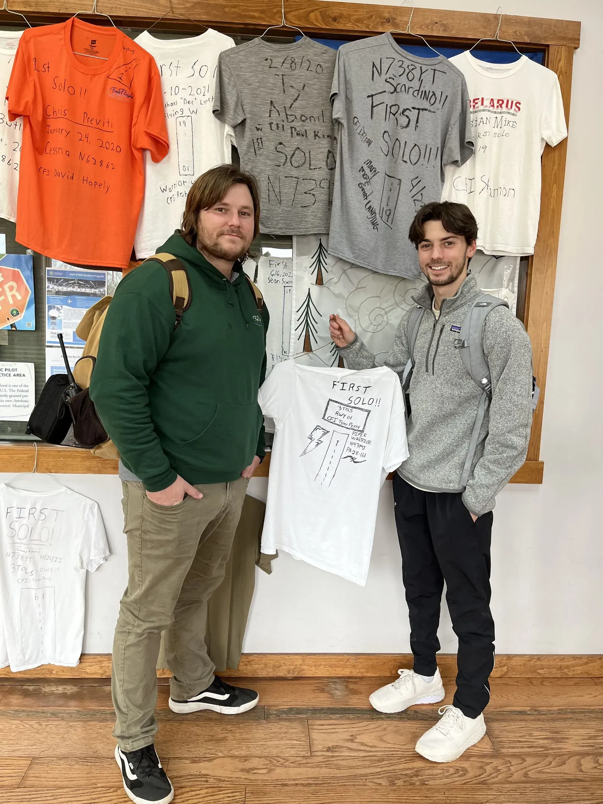 Two men posing with a white t-shirt in a shop. T-shirts with scribbled designs hang on the wall behind them.