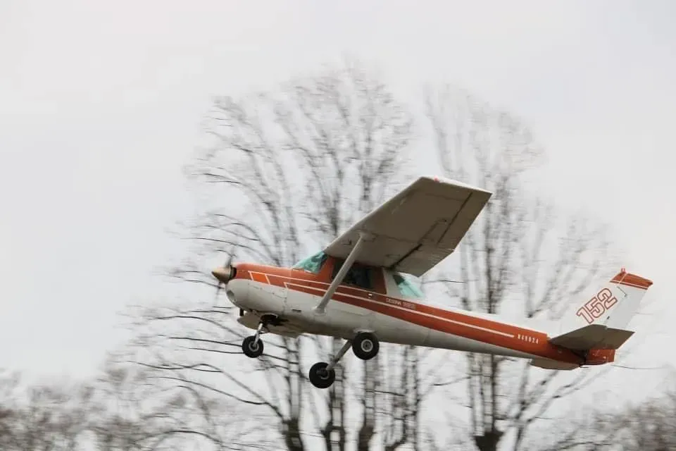 Orange and white single-engine airplane taking off against a cloudy sky and bare trees.