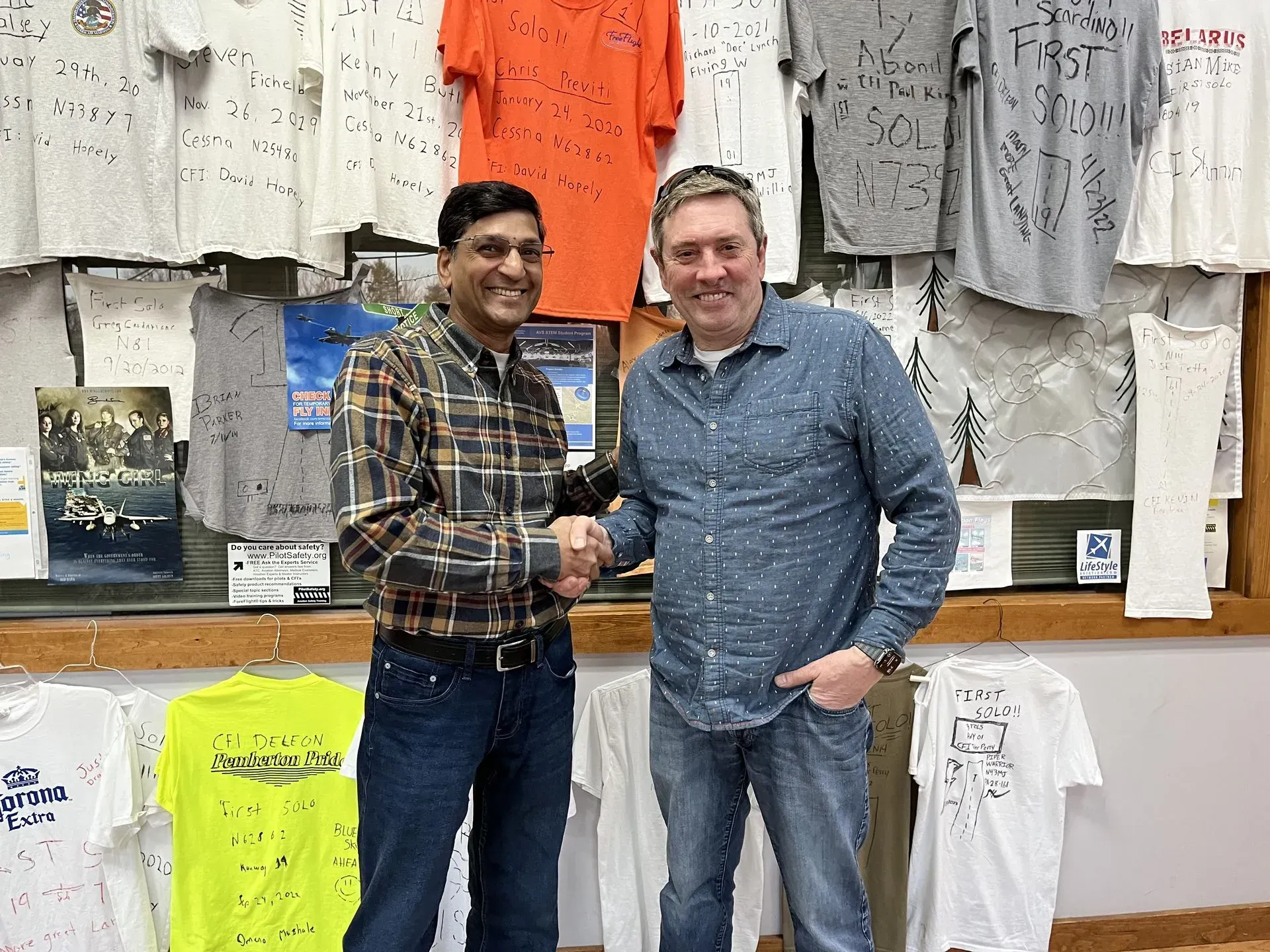 Two men shaking hands in front of a wall of t-shirts and signs. One man wears a plaid shirt, the other a blue button-down.