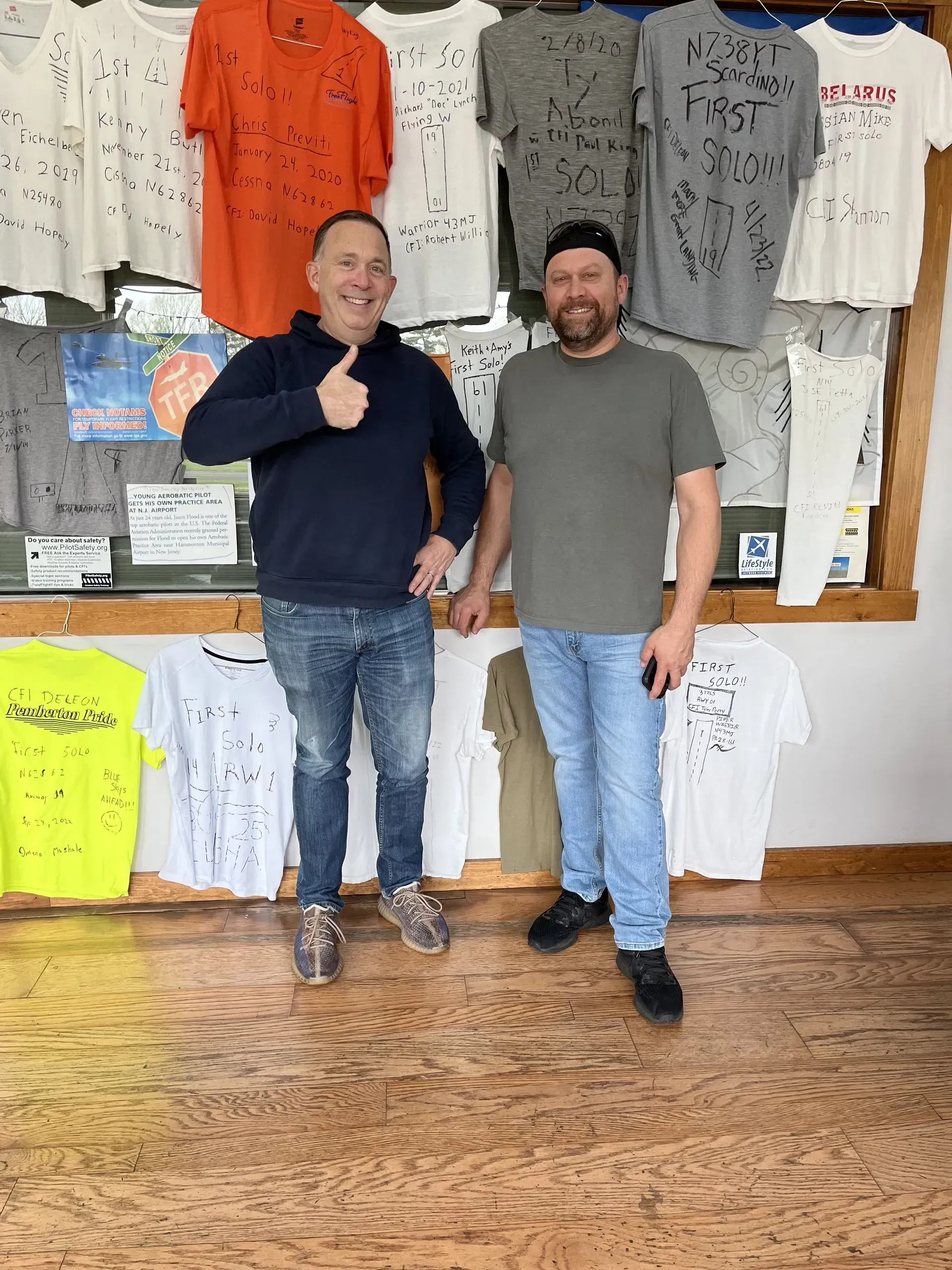 Two men pose in front of a wall of t-shirts. One gives a thumbs up. Setting appears to be a shop.