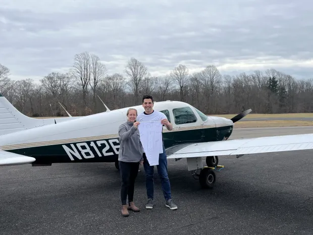 Couple holding baby onesie in front of small airplane; outdoors, overcast day.