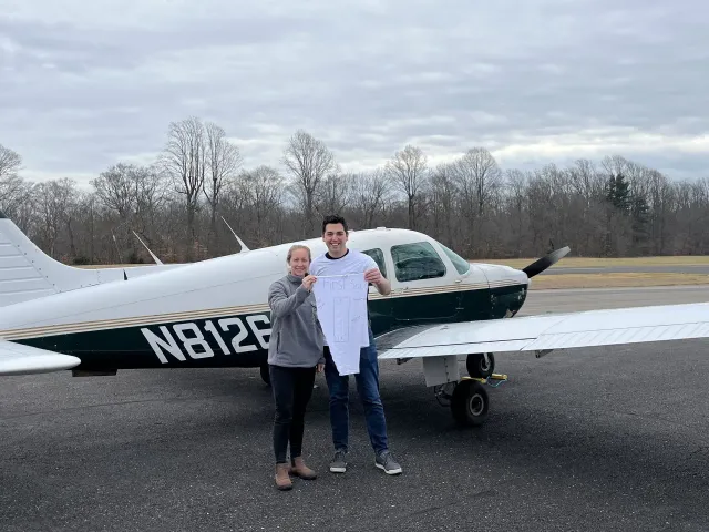 Couple holding baby onesie in front of small airplane; outdoors, overcast day.