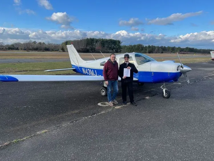 Two men stand next to a small, blue and white airplane on a runway. One holds a paper.