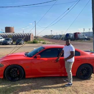 Man in white shirt stands next to a red Dodge Charger. Outdoors, sunny day, industrial setting.