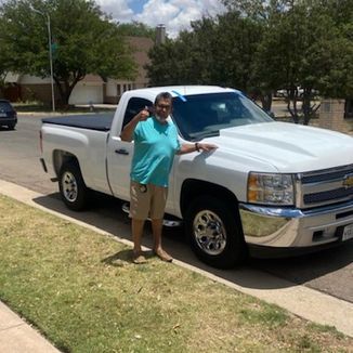 Man in blue shirt and khaki shorts stands beside a white Chevy pickup truck on a sunny day.
