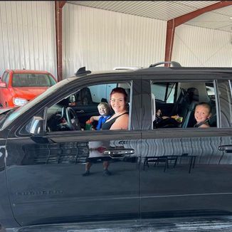 Woman and two children in a black SUV. Red truck in the background. Inside a garage.