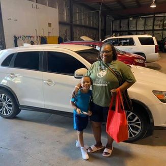 Woman and child standing next to a white car in a garage; woman holding a red bag, smiling.