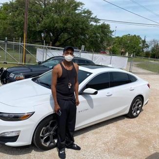 Man in black tank top and pants stands by a white Malibu car with tinted windows, outdoors.