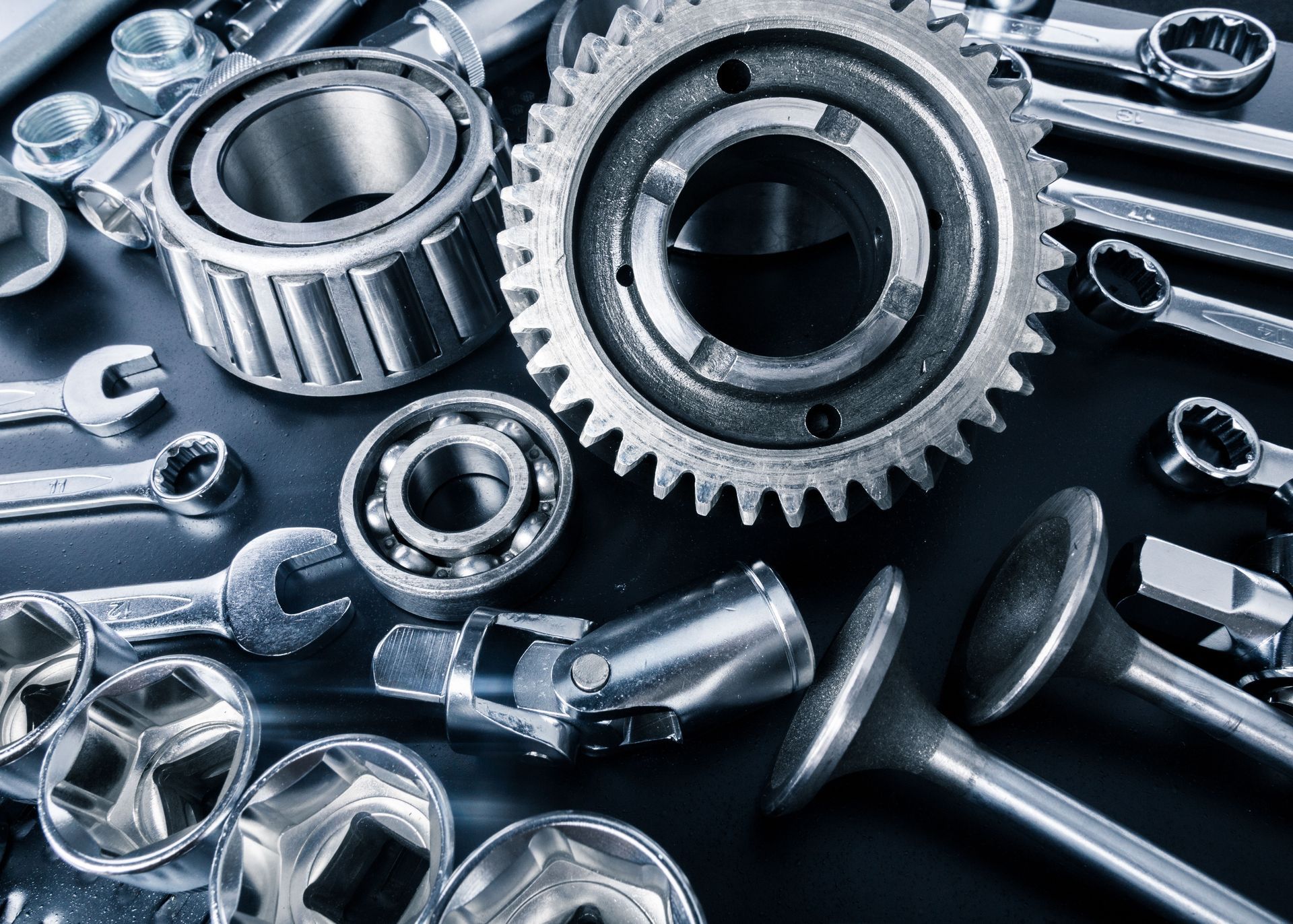 Close-up of assorted metal mechanical parts, including gears, bearings, and wrenches, on a dark surface.