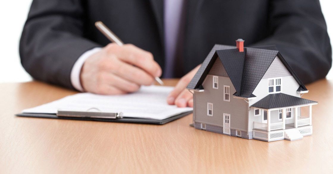 A man in a suit is writing on a clipboard next to a model house.