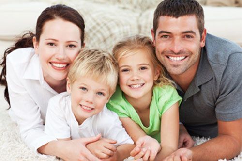 Family of four smiling and lying down together on a light-colored rug.