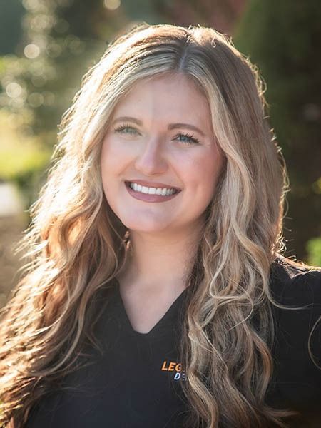 Woman with long wavy blonde hair smiling outdoors in front of greenery.