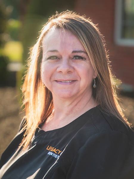 Woman with long hair smiles in outdoor setting, wearing a black top with logo.