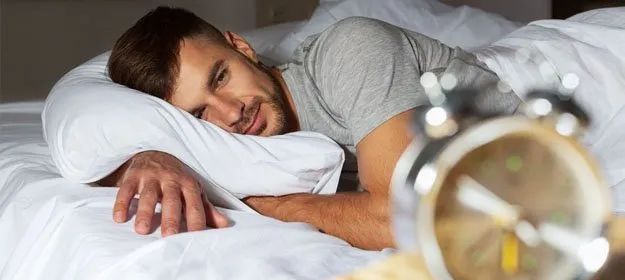 A man is sleeping in bed next to an alarm clock.