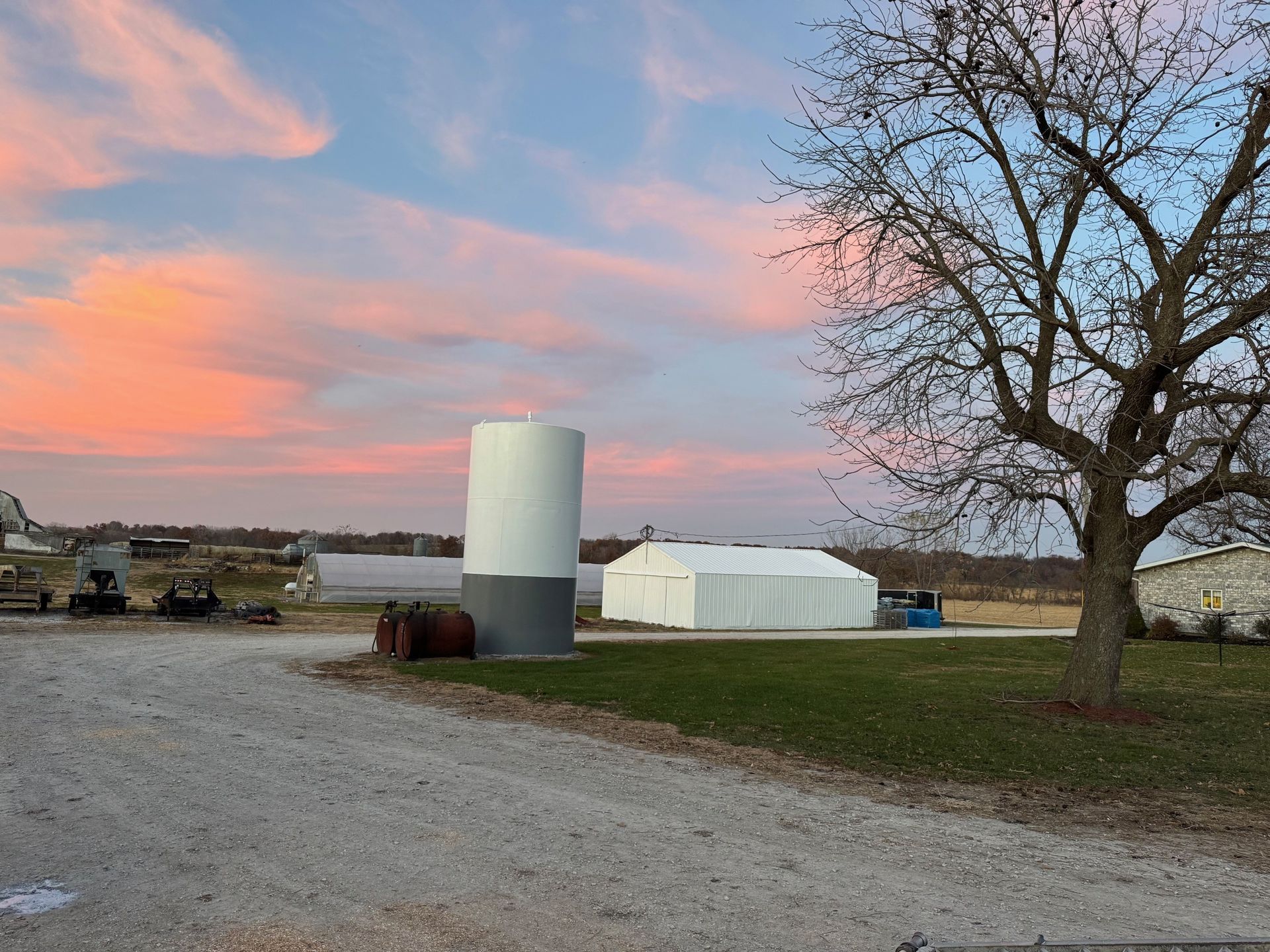 Rural farmyard at sunset with grain silo, white barns, gravel drive, and bare tree under pink clouds
